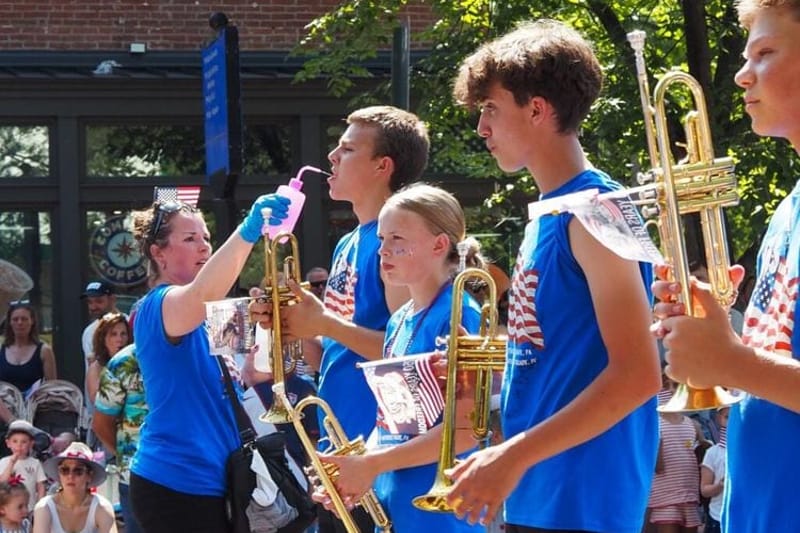 A chaperone from Nebraska squirted water into the mouth of a Grand Island Senior High School marching band member at the Capitol Hill Fourth of July Parade on Saturday, July 4, 2026 in Washington, D.C. The band marched in the smaller parade after the National Independence Day Parade was canceled due to the extreme heat. (Photo by Ashley Murray/States Newsroom)