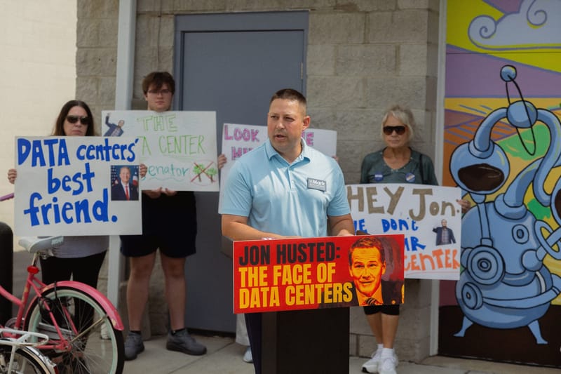 Wood County auditor candidate Mark Wongrowski speaks at a Bowling Green rally this week as residents protest U.S. Sen. Jon Husted’s record of promoting data center development in Ohio. Behind him, demonstrators hold signs criticizing Husted as Meta builds an $800 million data center near Bowling Green. (Photo: X)